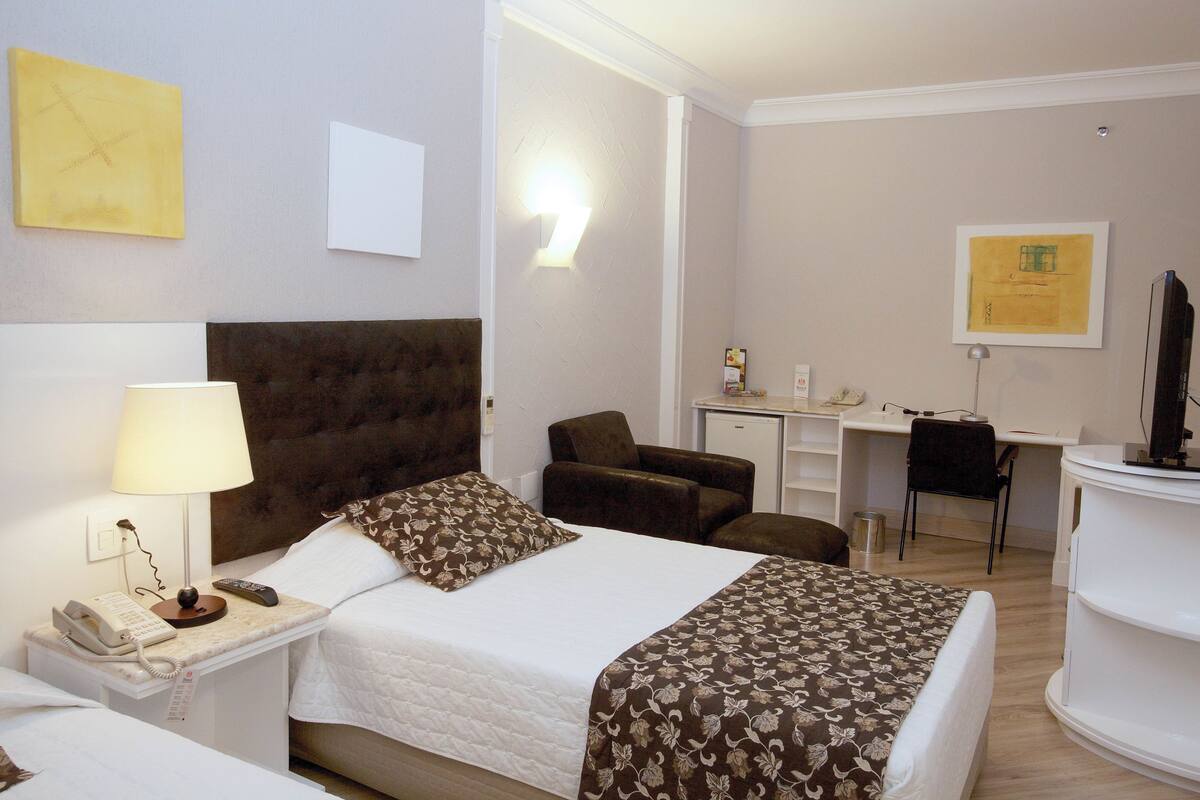 Hotellihuone, jossa on sänky, yöpöytä, lamppu ja työpöytä tuolilla.
