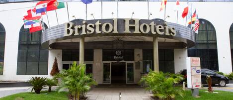 The exterior of the Bristol Hoteliers building with multiple flags, a sign indicating the entrance for vehicles, and a paved entrance area with potted plants.