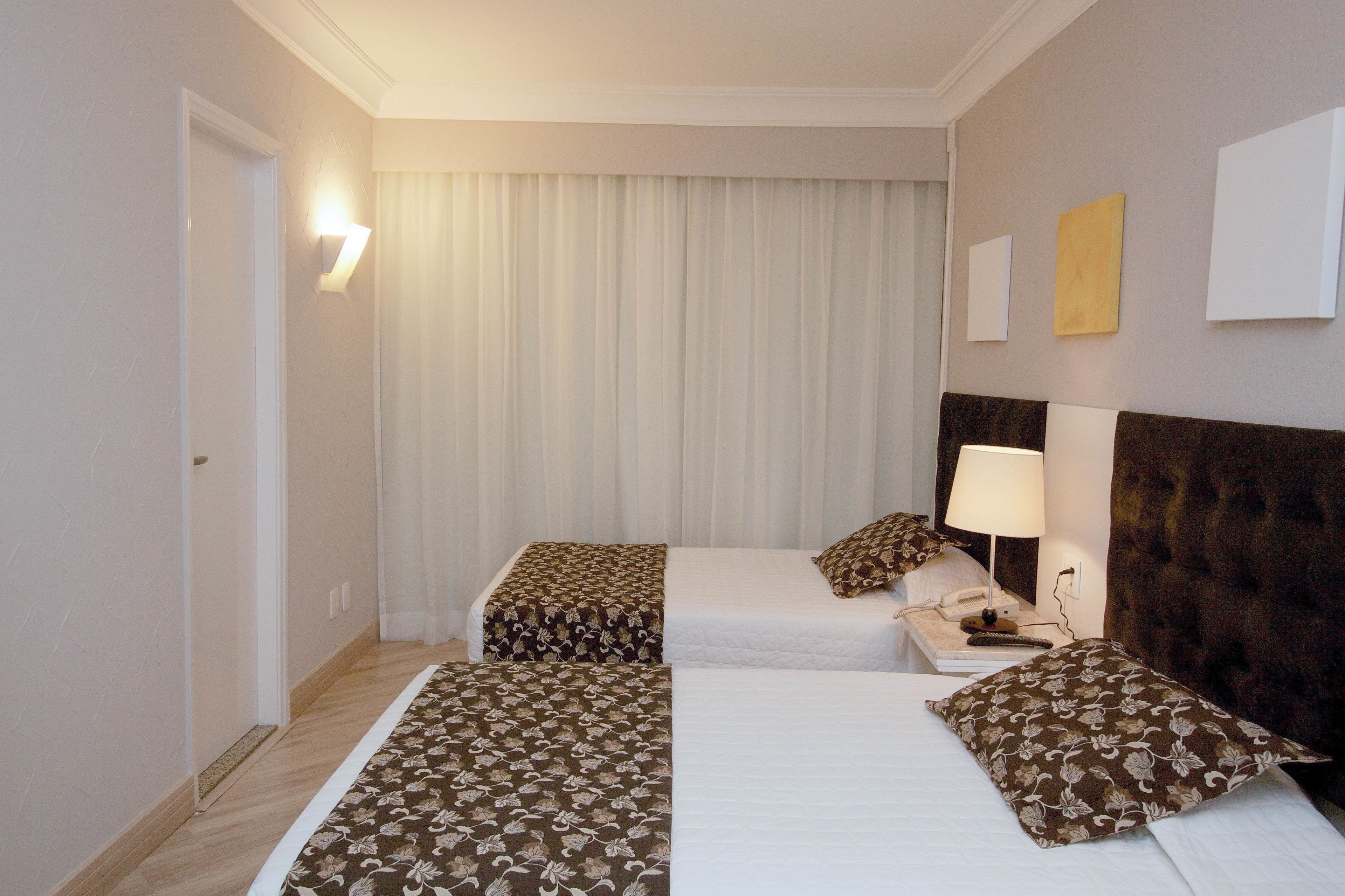 Quarto de hotel com duas camas, uma mesa de cabeceira com luminária, telefone e iluminação na parede.