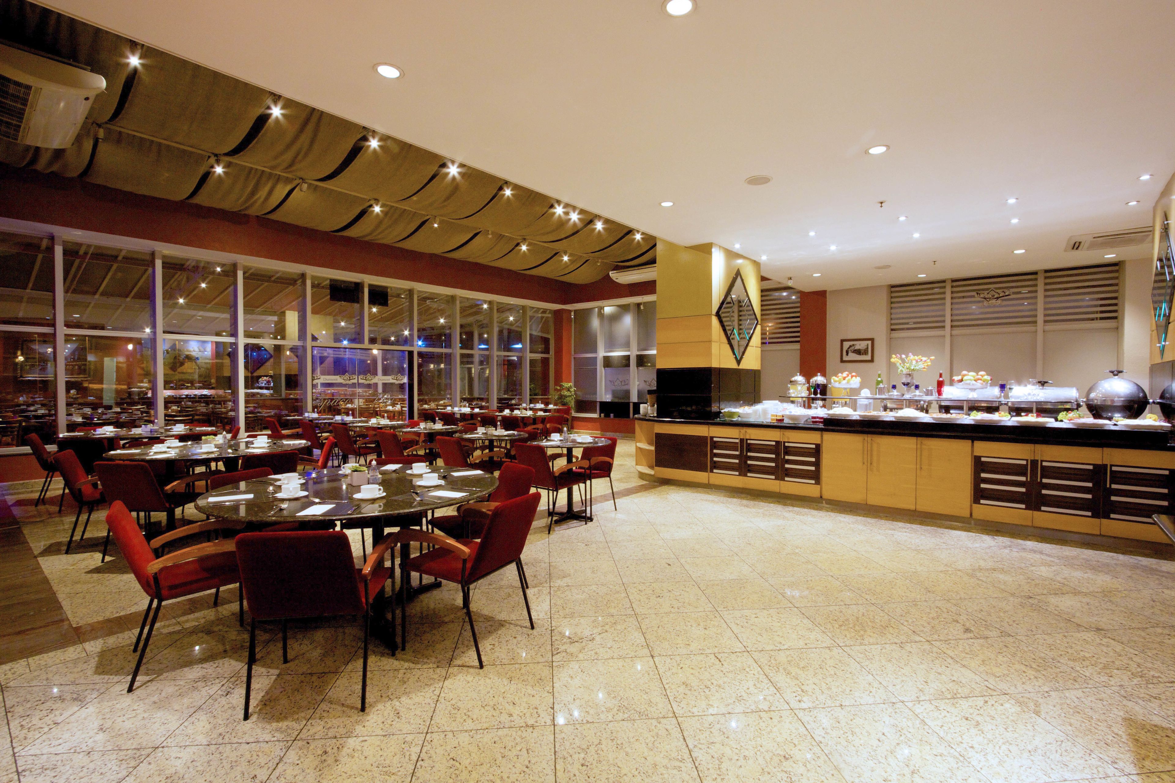 Ampla área de refeições com várias mesas e cadeiras, um buffet e grandes janelas que permitem entrada de luz natural.