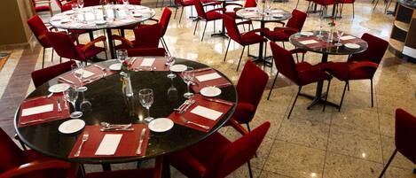 Un restaurante con varias mesas redondas y sillas rojas, dispuesto en un espacio amplio con piso de baldosas.