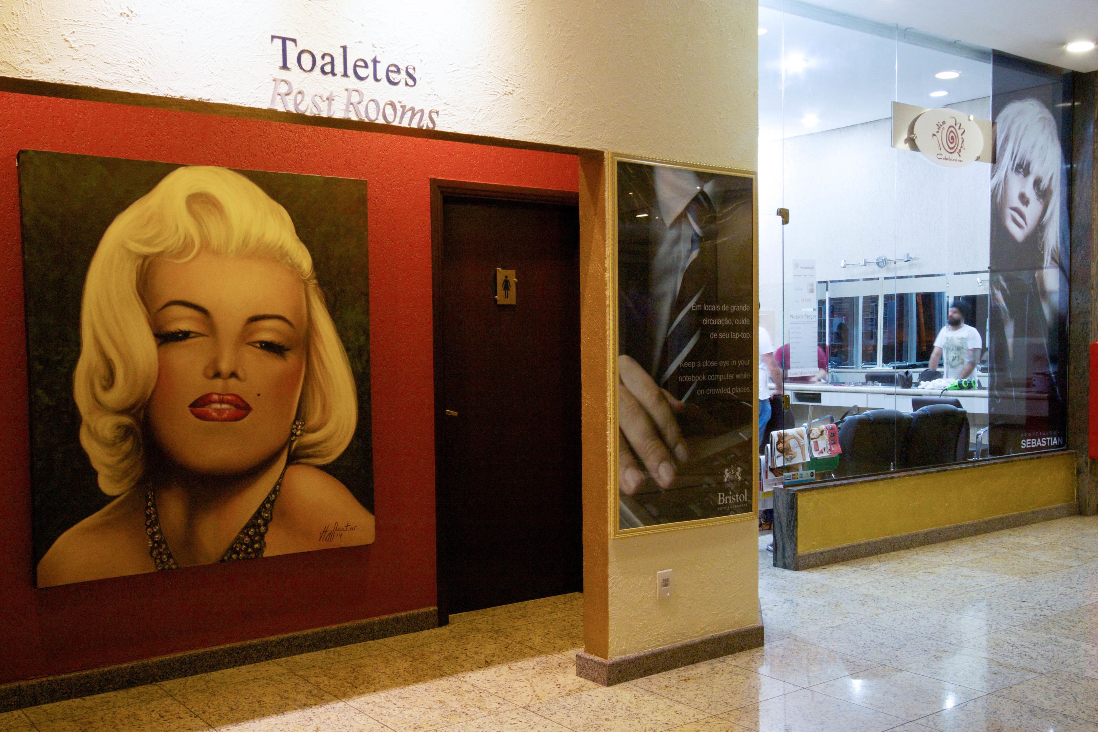 Corredor de hotel com placa de banheiro, uma porta e um grande cartaz de uma mulher.