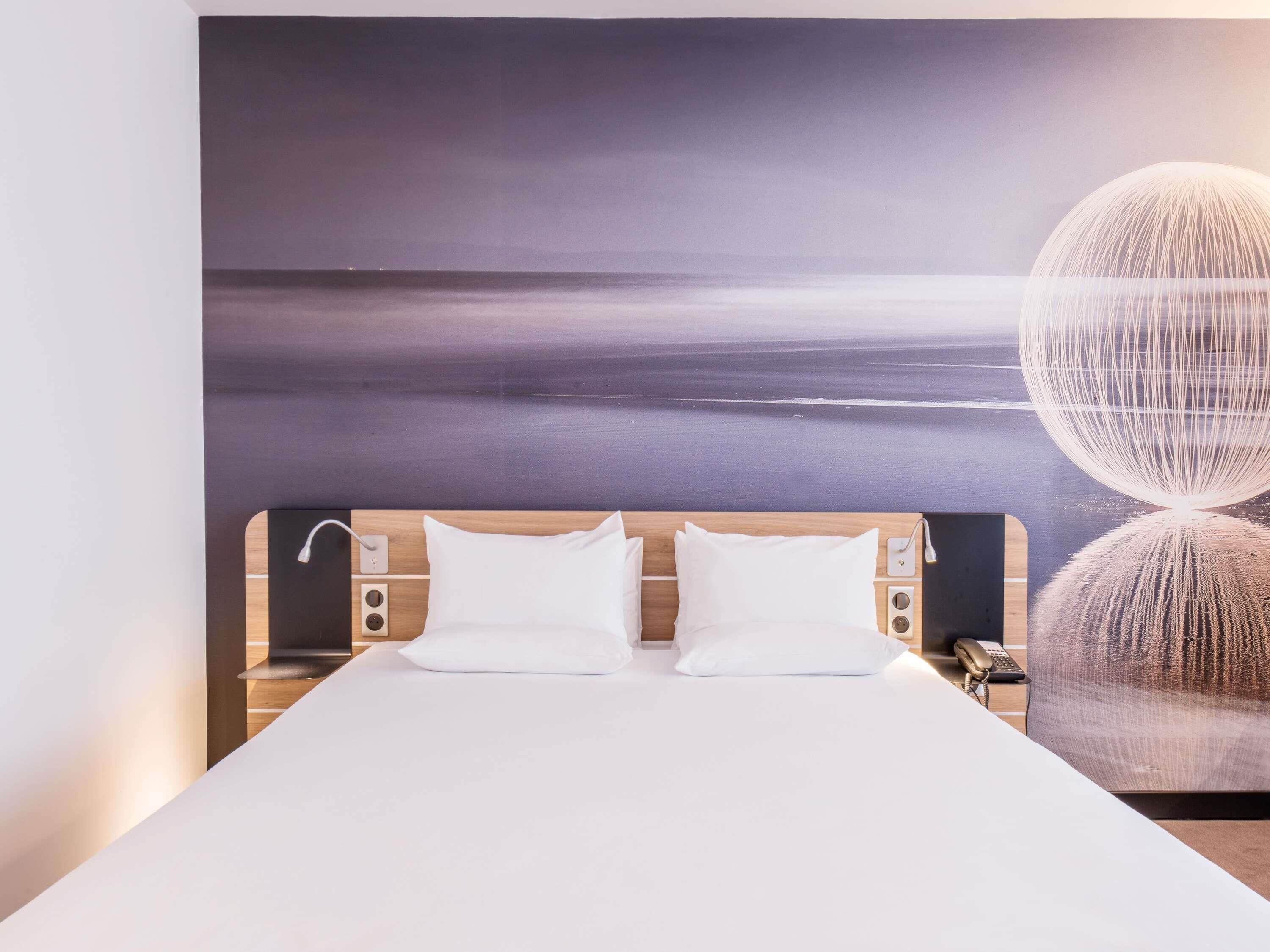 A modern hotel room with a large bed, a headboard with built-in lighting, a wall-mounted phone, and a large window with a view of the sea.