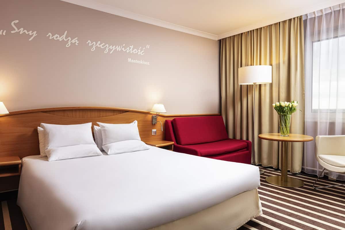 Ein Hotelzimmer mit einem großen Bett, einem roten Sessel, einem kleinen Tisch mit einer Vase und einer Wand mit einem Zitat.