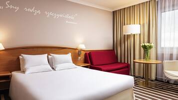 A hotel room with a large bed, a red armchair, a small table with a vase, and a wall with a quote.