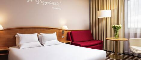 A hotel room with a large bed, a red armchair, a small table with a vase, and a wall with a quote.