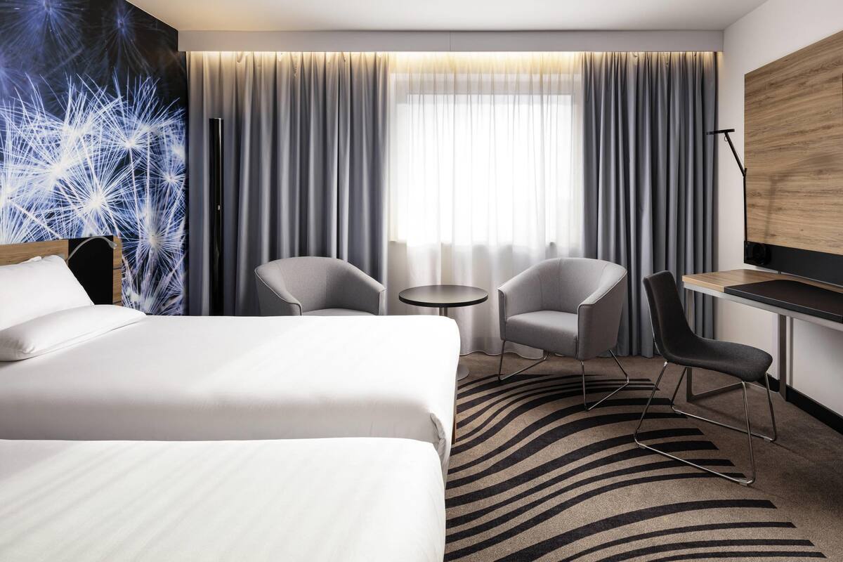 Ein Hotelzimmer mit einem Bett, zwei Sesseln, einem kleinen Tisch, einem Schreibtisch und einem großen Fenster mit Vorhängen.
