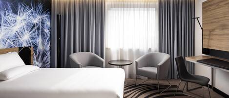 A hotel room with a bed, two armchairs, a small table, a desk, and a large window with curtains.