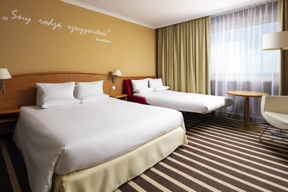Ein Hotelzimmer mit zwei Betten, einem roten Sessel, einem kleinen Tisch und einer Wand mit einem Zitat.