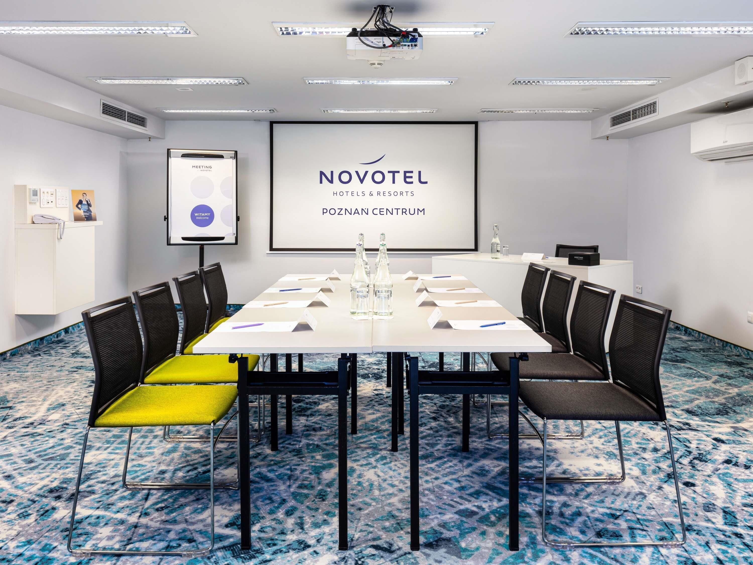 A meeting room with a white table, black chairs, and a projector screen displaying 'NOVOTEL HOTELS & RESORTS POZNAN CENTRUM'.