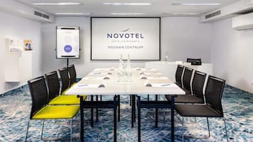 A meeting room with a white table, black chairs, and a projector screen displaying 'NOVOTEL HOTELS & RESORTS POZNAN CENTRUM'.