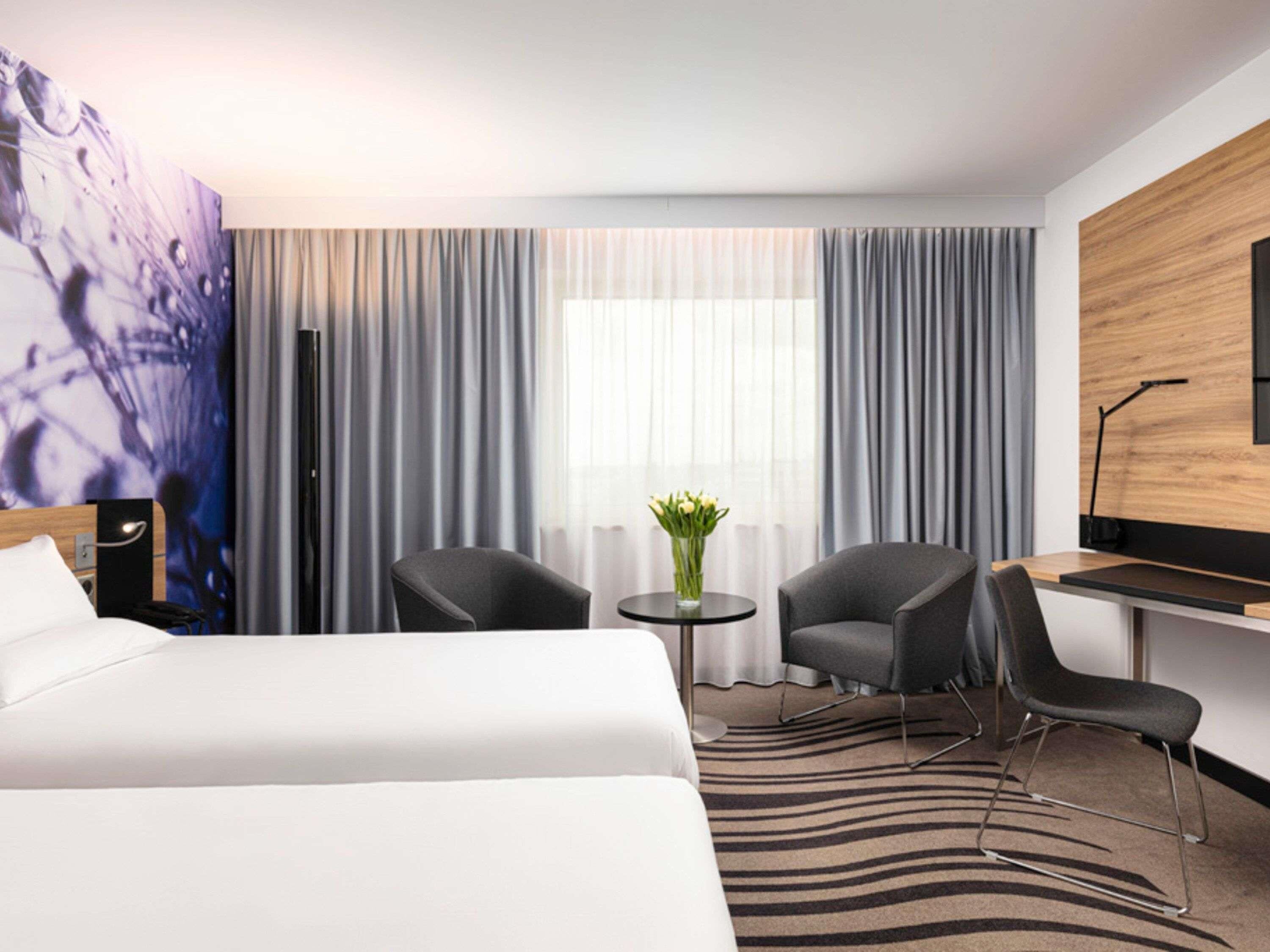 A hotel room with two beds, a desk, a chair, a small table with a vase of flowers, and a large abstract painting on the wall.