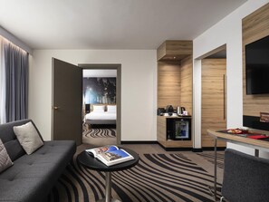 Una habitación de hotel moderna con cama, una zona de estar con una revista, una kitchenette pequeña y un televisor.