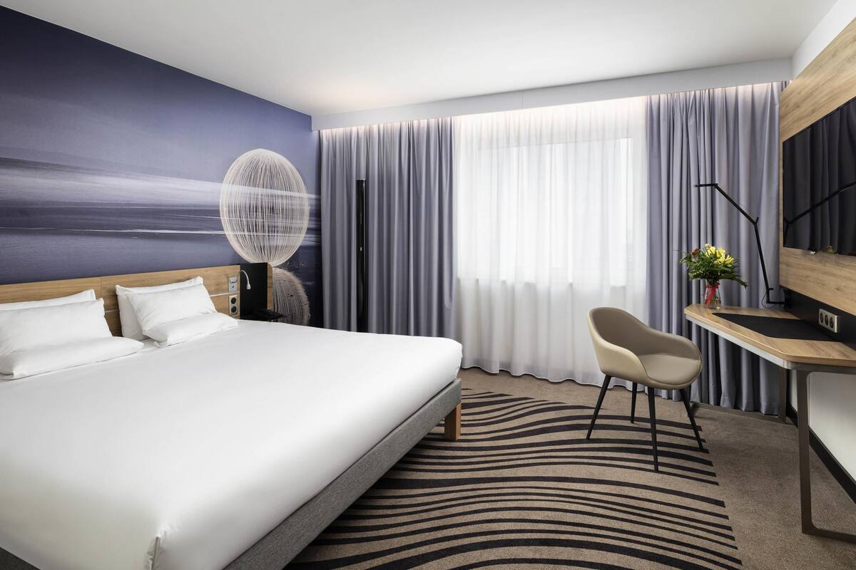 Ein Hotelzimmer mit einem großen Bett, einem Schreibtisch, einem Stuhl, einem großen Fenster mit Vorhängen und einem Wandbild.