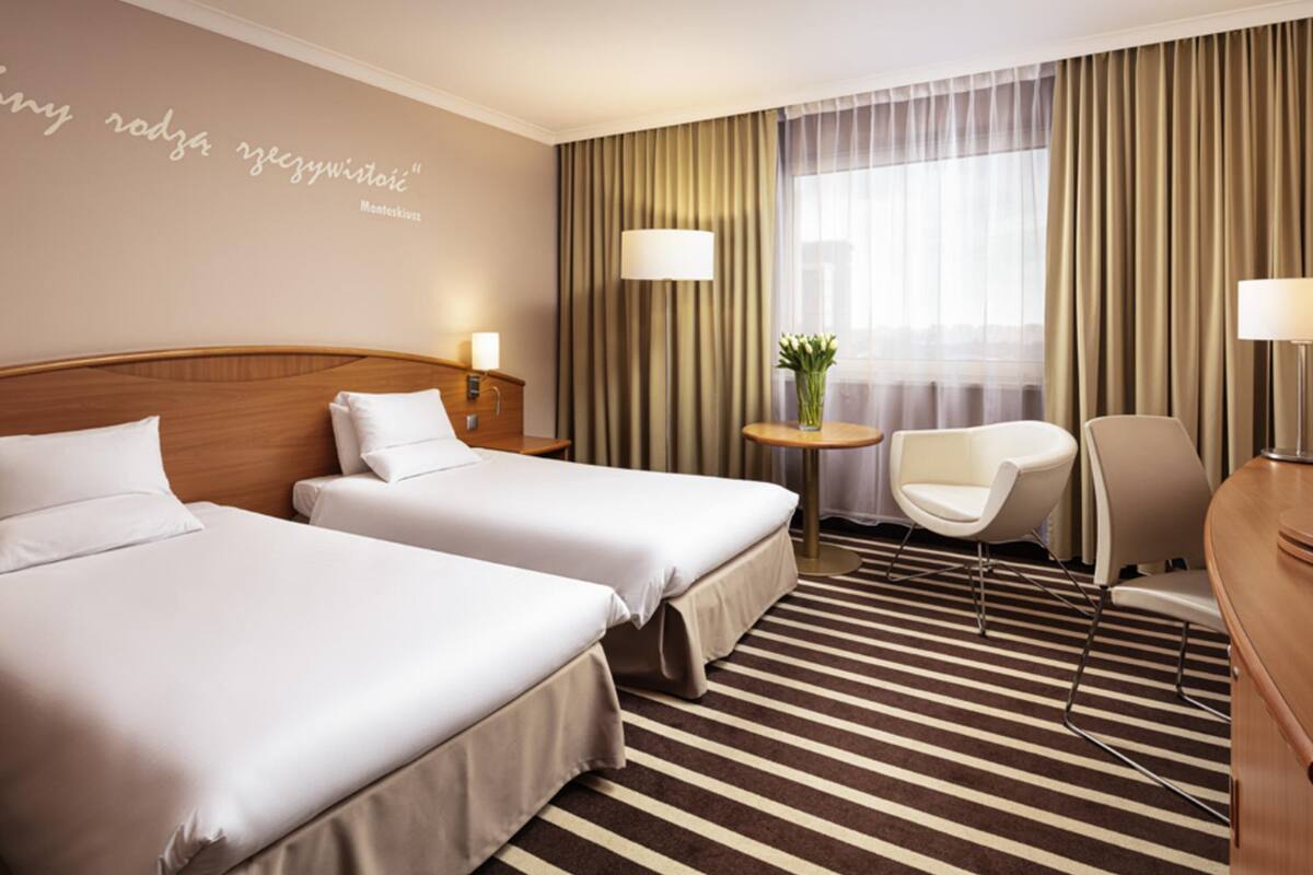 Ein Hotelzimmer mit zwei Betten, einem Schreibtisch, einem Stuhl, einem kleinen Tisch mit Blumenvase und einem Fenster mit Vorhängen.