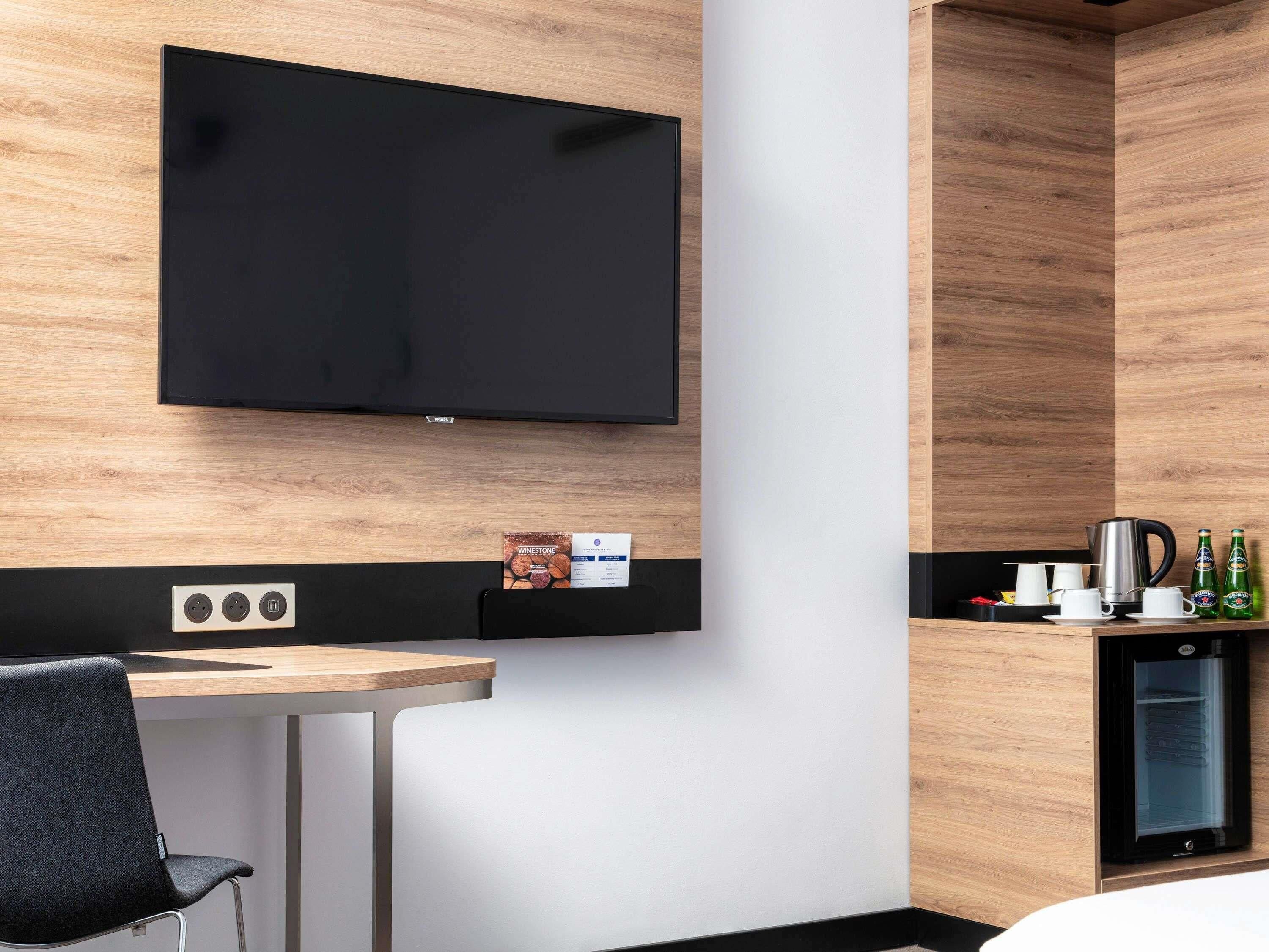 A modern hotel room with a flat-screen TV mounted on the wall, a small kitchenette with a coffee maker, and a compact refrigerator.