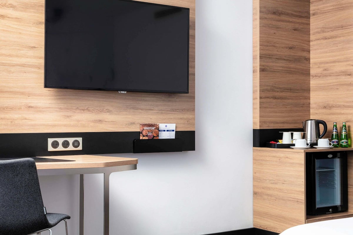 A modern hotel room with a flat-screen TV mounted on the wall, a small kitchenette with a coffee maker, and a compact refrigerator.