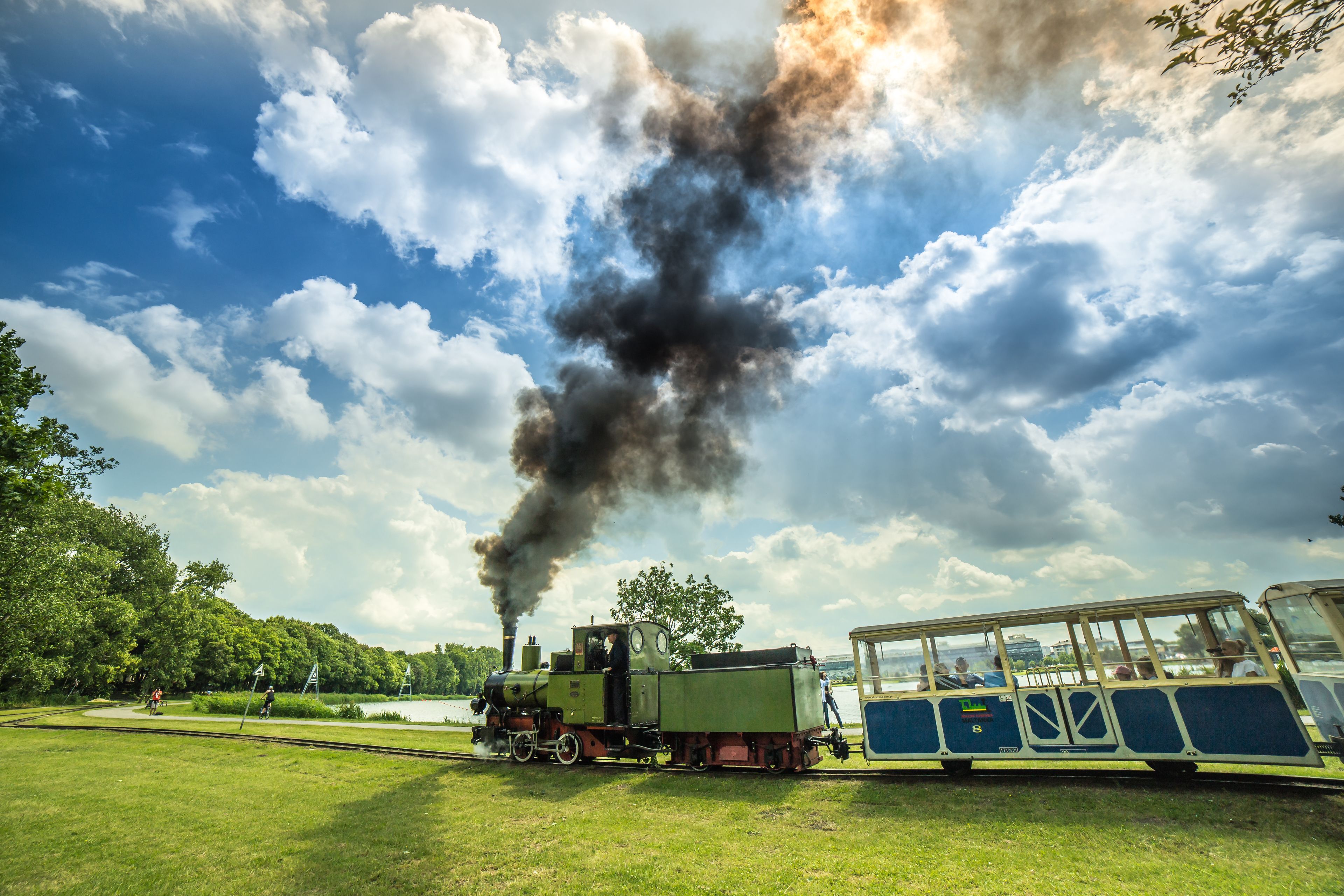 A steam locomotive emitting smoke, a passenger train, and a park setting with trees and a lake.