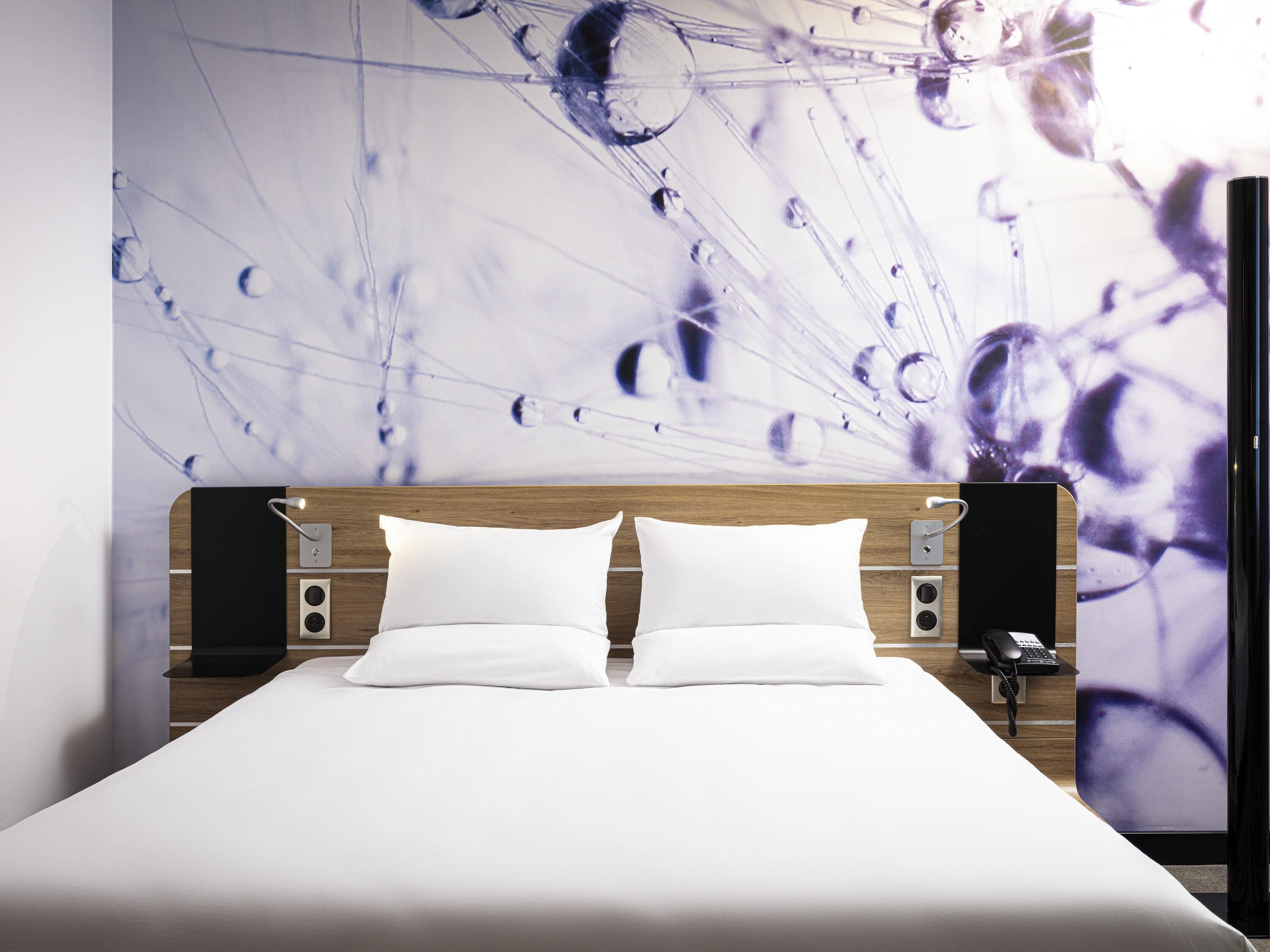 A hotel room with a bed, two white pillows, a wooden headboard, a wall-mounted phone, and a wall art depicting abstract shapes.