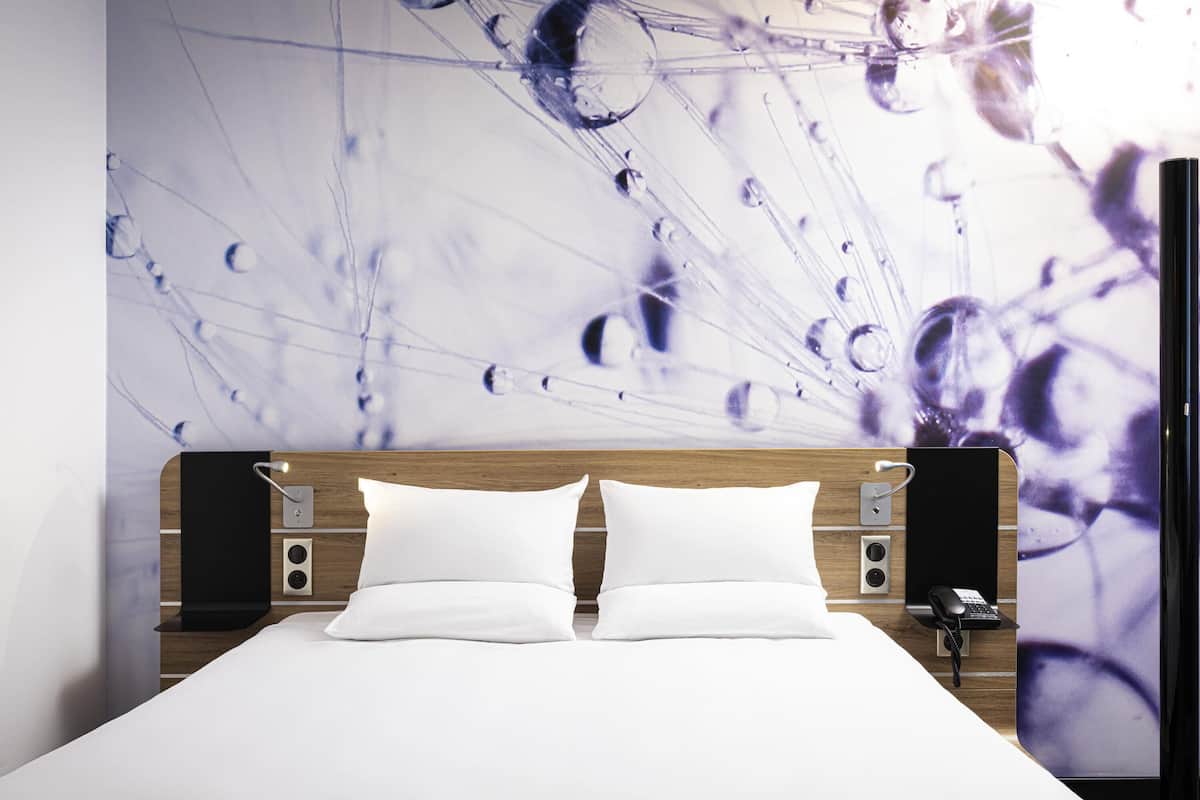 A hotel room with a bed, two white pillows, a wooden headboard, a wall-mounted phone, and a wall art depicting abstract shapes.