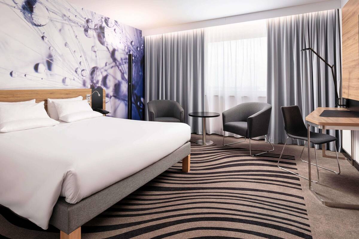 Ein Hotelzimmer mit einem großen Bett, zwei Sesseln, einem kleinen Tisch und einem Schreibtisch mit Stuhl.