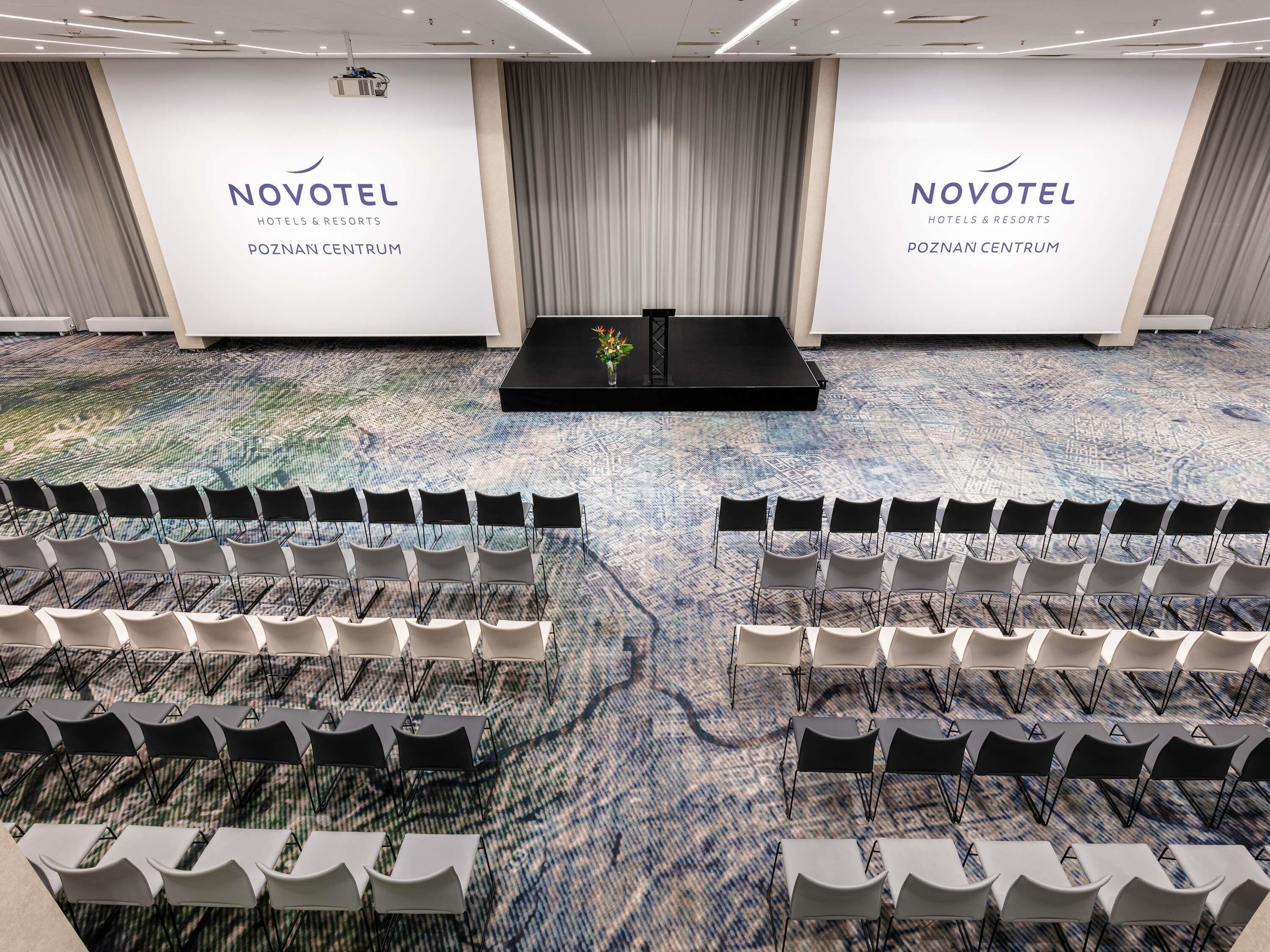 Un salón de conferencias con filas de sillas y un escenario, marca NOVOTEL, y suelo alfombrado.