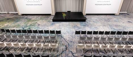 Un salón de conferencias con filas de sillas y un escenario, marca NOVOTEL, y suelo alfombrado.