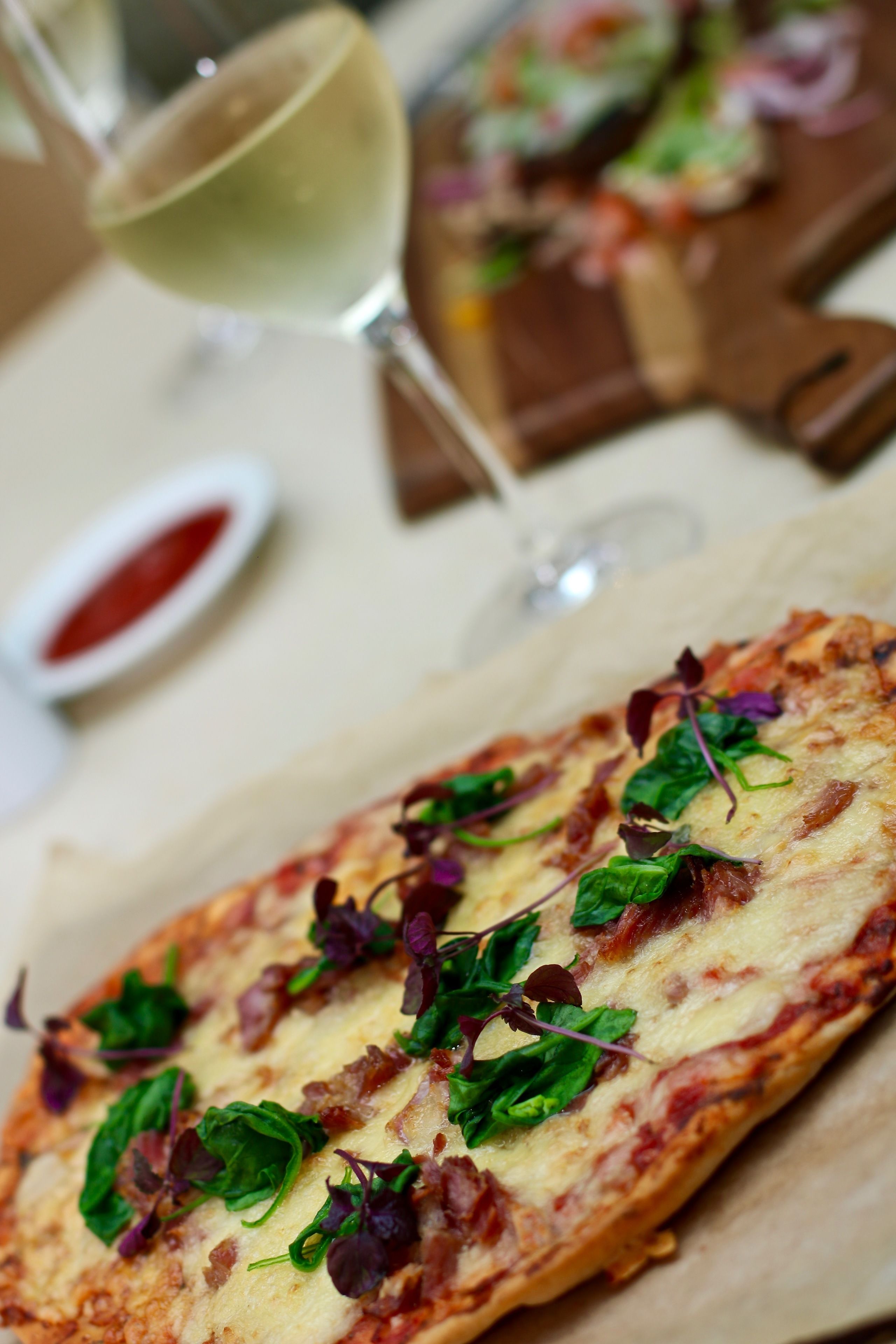 A pizza with greens and cheese, a glass of white wine, and a small bowl of sauce.