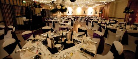 Un salón de banquetes con mesas redondas dispuestas para un evento formal, destacando manteles blancos, cubertería y cristalería.