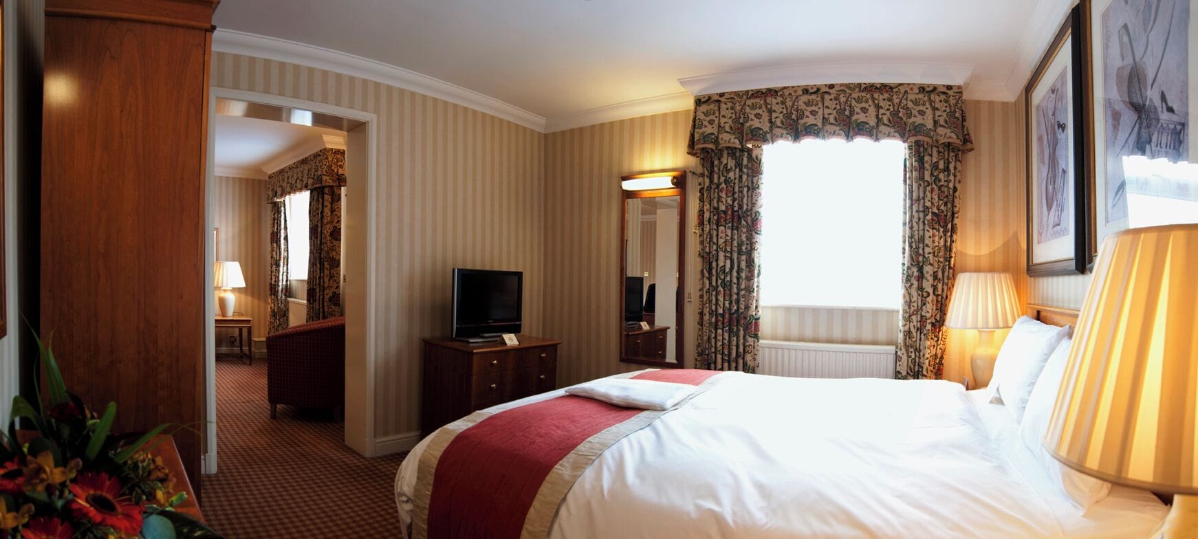 Quarto de hotel com uma cama grande, televisão, escrivaninha com luminária e janela com cortinas.