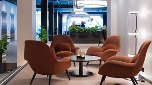 Un área de descanso moderna con sillones naranjas alrededor de una mesa redonda, una lámpara colgante de cristal transparente y un mostrador de recepción al fondo.