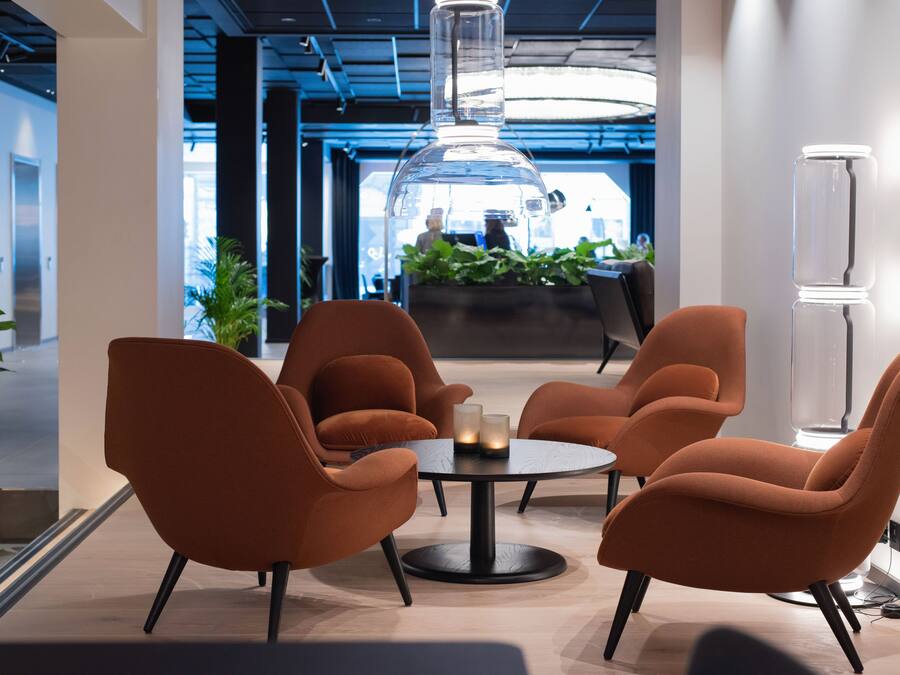 Un área de descanso moderna con sillones naranjas alrededor de una mesa redonda, una lámpara colgante de cristal transparente y un mostrador de recepción al fondo.