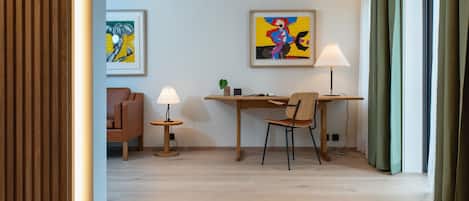 A modern hotel room with a wooden floor, a desk with two chairs, a lamp, and a framed painting on the wall.