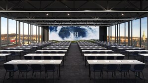 Un salón de conferencias con filas de mesas y sillas, ventanas amplias y vistas a un paisaje urbano.