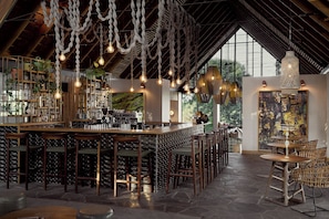 Un espace bar doté d’un plafond haut, de luminaires suspendus et offrant une vue sur l’extérieur grâce à de grandes fenêtres.