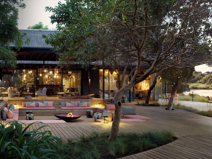 An outdoor seating area with a fire pit, sofas, and tables, surrounded by a wooden deck and greenery.