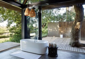 A modern outdoor bathroom with a freestanding bathtub, wooden deck, and a large window overlooking a natural landscape.