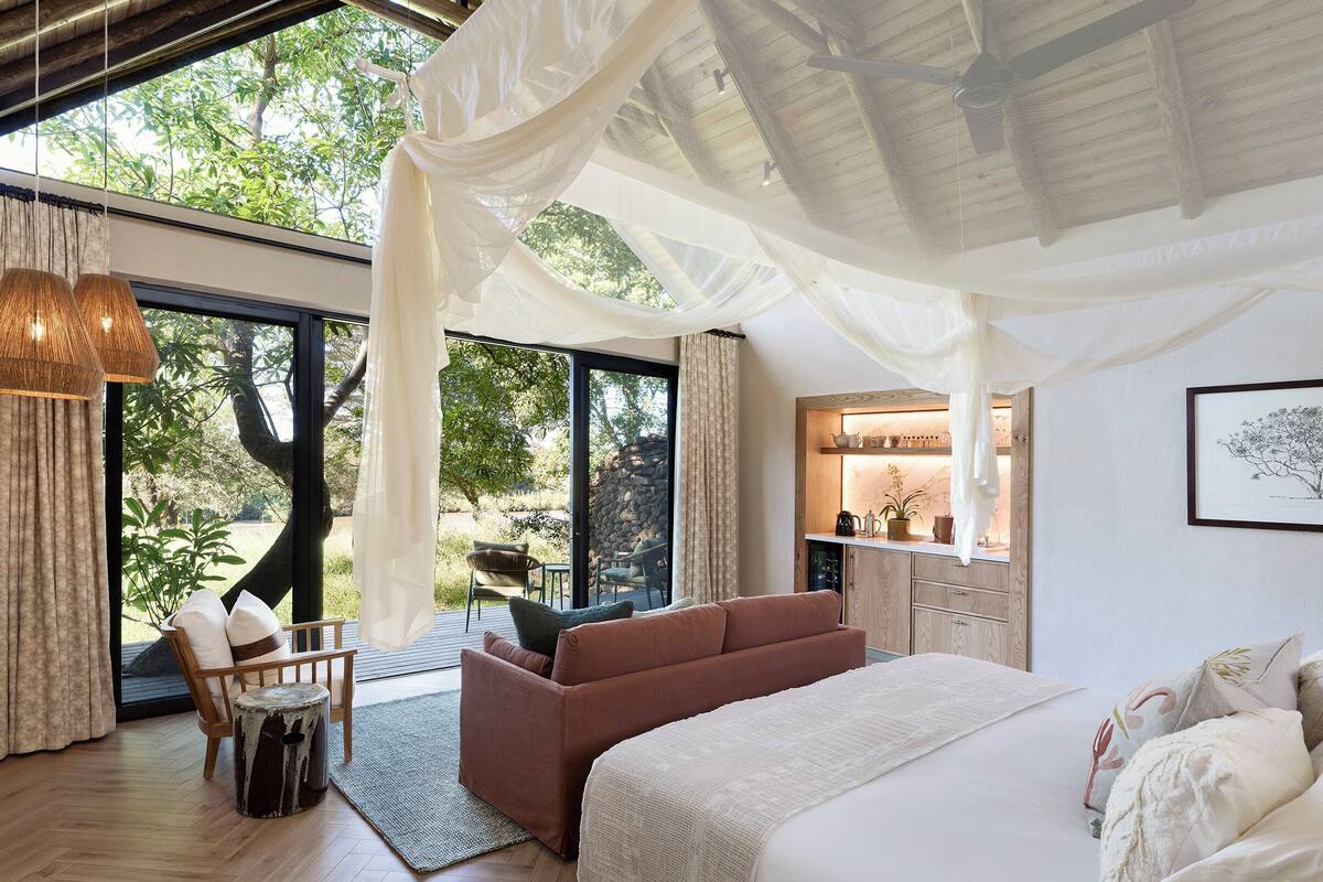 Een slaapkamer met een groot bed, een bank, een stoel en een kleine tafel. Er is een raam met uitzicht op bomen en een balkon.