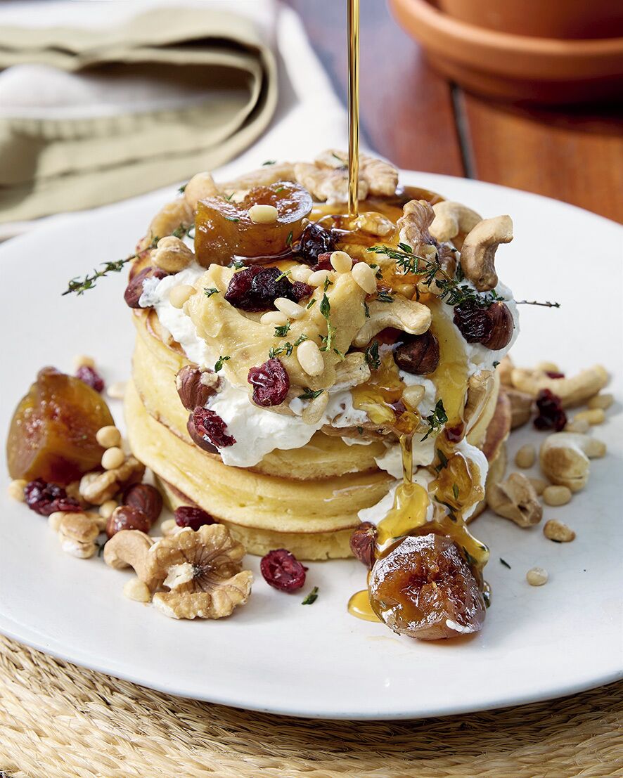 A stack of pancakes with syrup, nuts, and dried fruits on a white plate.