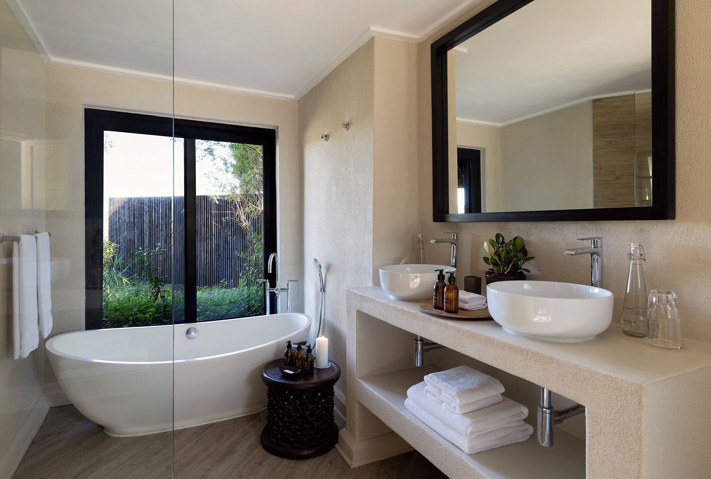 A modern bathroom with a freestanding bathtub, two sinks, a large mirror, and a view of greenery outside.