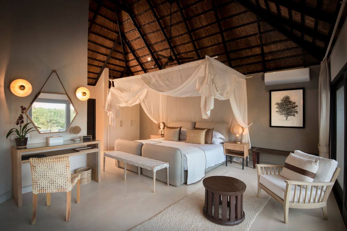 Een slaapkamer met een hemelbed, een bureau, een stoel, een bankje en een klein rond tafeltje.