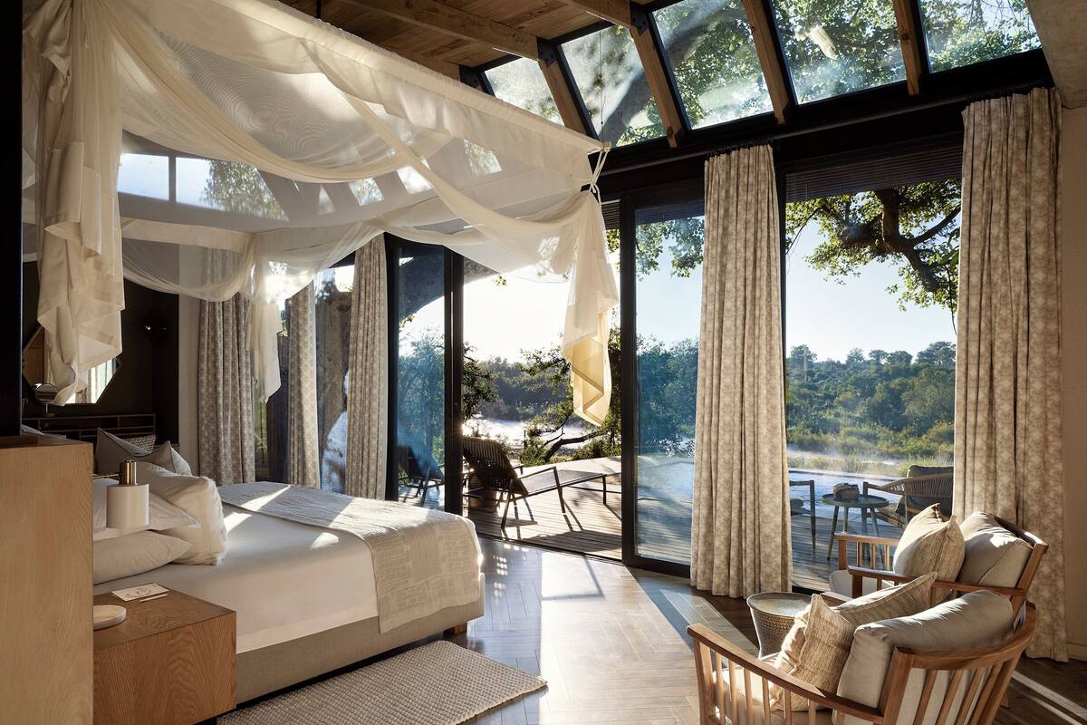 Een slaapkamer met een groot bed, een houten stoel, een klein tafeltje en een uitzicht op een natuurlandschap door grote ramen.