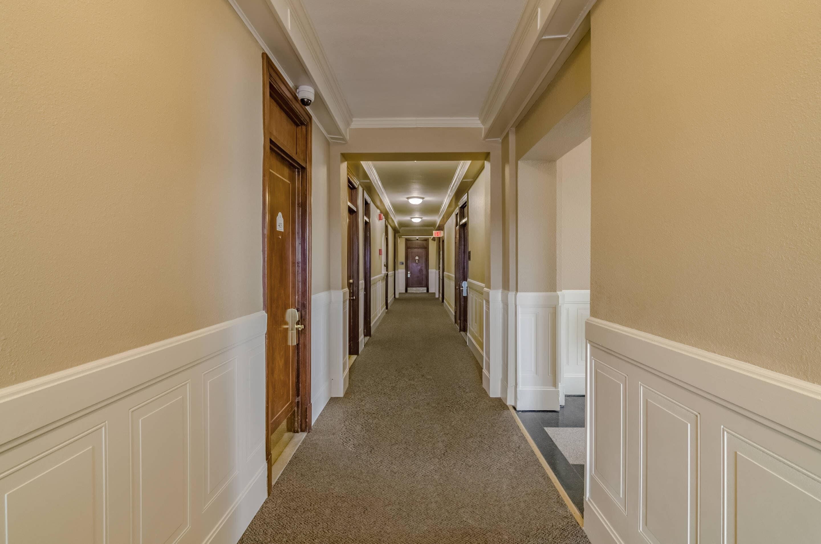 A hotel hallway with a carpeted floor, white wainscoting, and wooden doors.