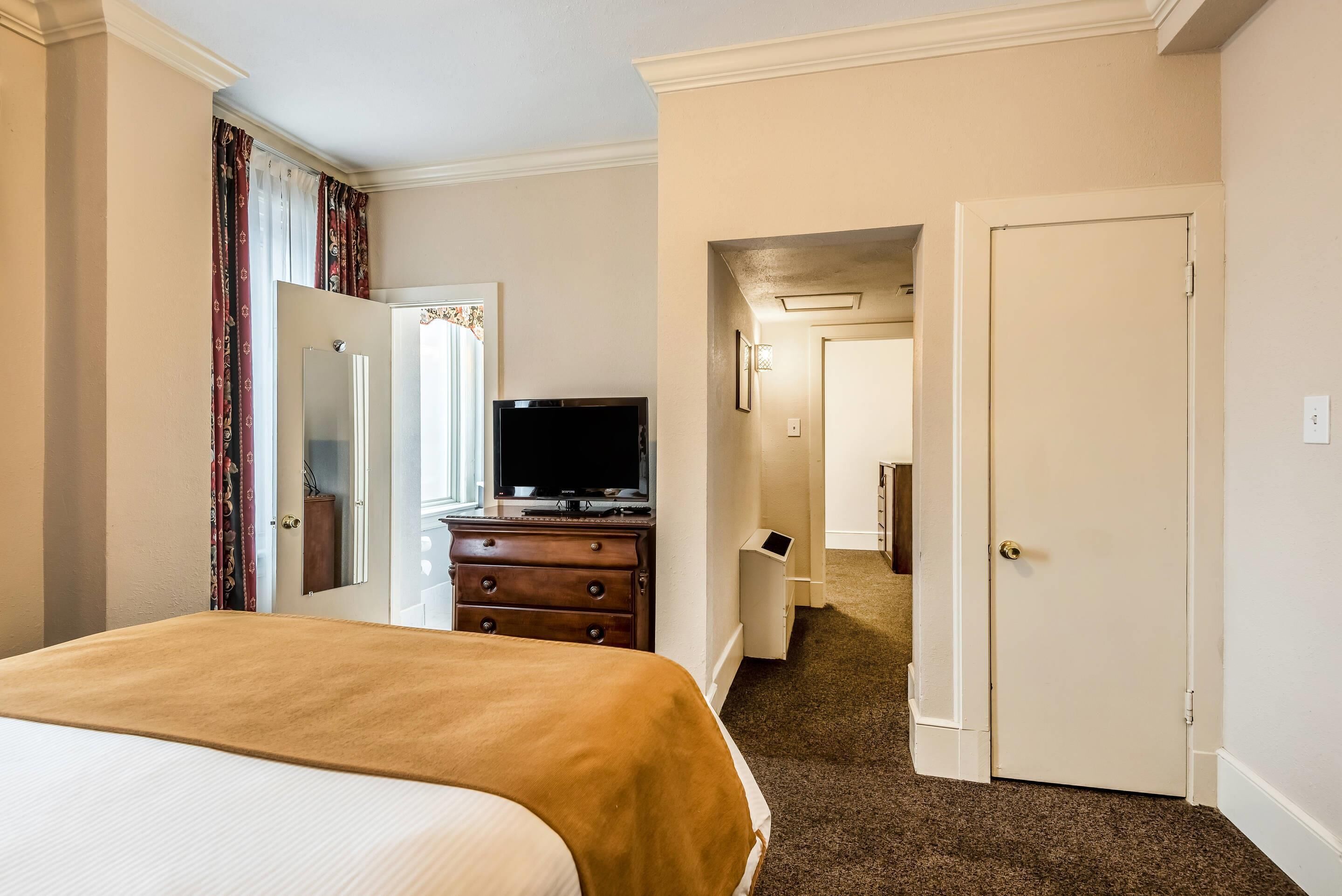 A hotel room with a bed, a dresser, a television, and a door leading to another room.