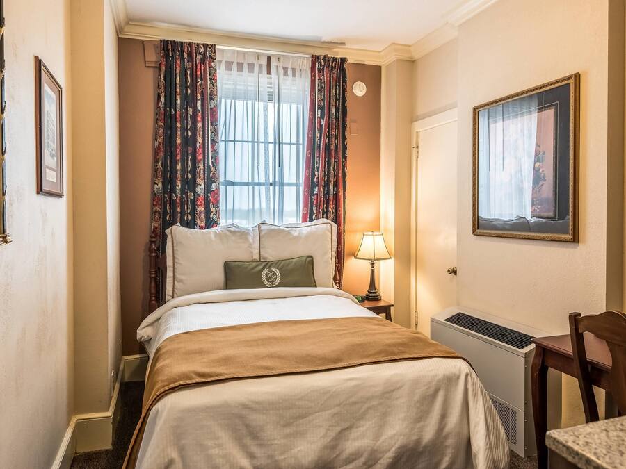 Quarto de hotel com cama, escrivaninha, cadeira, janela com cortinas, radiador e um quadro na parede.