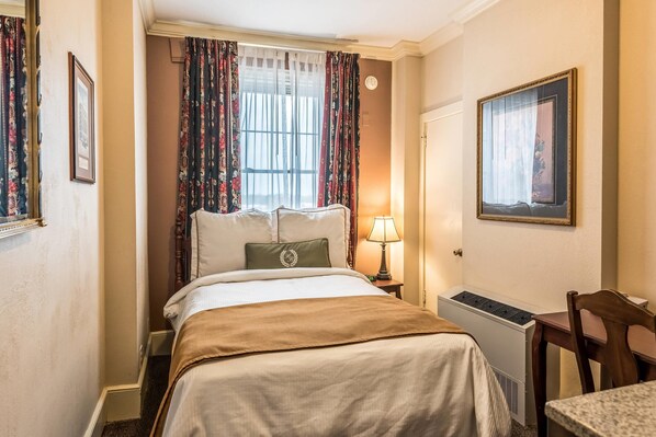 A hotel room with a bed, a desk, a chair, a window with curtains, a radiator, and a framed picture on the wall.