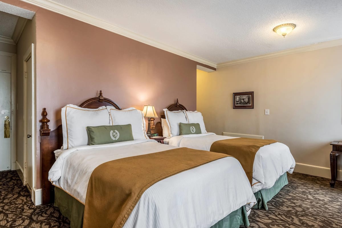 Habitación de hotel con dos camas, cada una con cabecero de madera, ropa de cama blanca y almohadas verdes. Hay una mesita de noche con una lámpara, un cuadro enmarcado en la pared y suelo alfombrado.