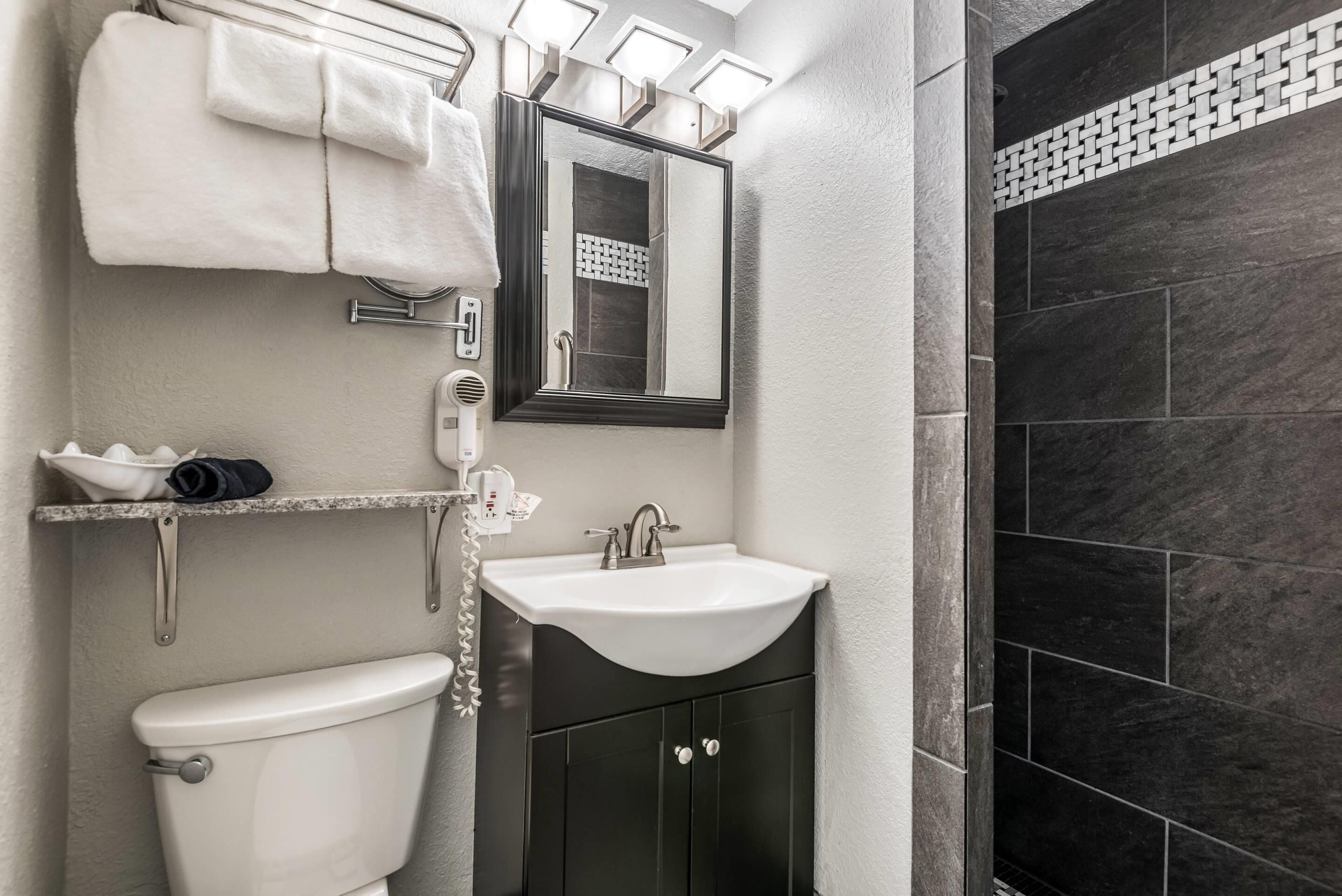 A modern bathroom with a white toilet, a sink, and a dark cabinet. There is a towel rack with towels, a mirror, and a shower area with dark tiles.