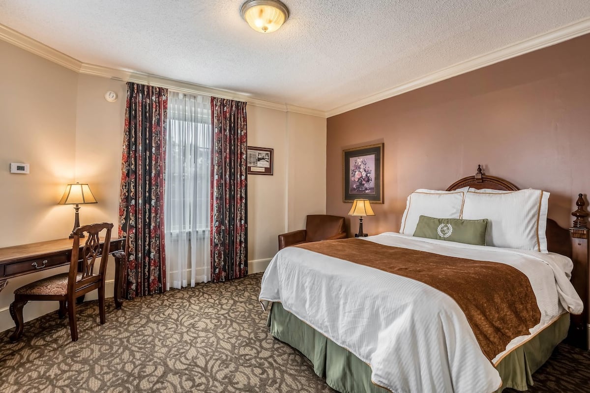 Una habitación de hotel con cama, escritorio, silla y ventana con cortinas.