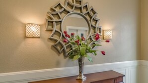 A wall with a decorative mirror, two wall sconces, and a vase with red flowers on a wooden console table.